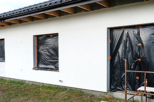 A newly applied layer of white silicone plaster on the wall of the house, visible metal scaffolding and boards, window is secured with black foil.